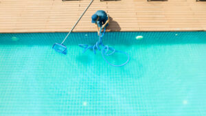 Man in blue uniform cleaning a swimming pool with a long net on a pole. The water is clear and tile-lined, with a wooden deck surrounding it.